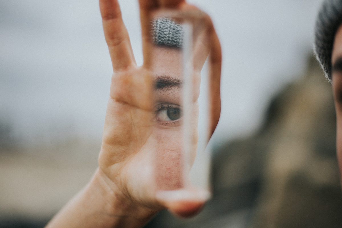 Photograph of a White person holding a fragmentated shard of mirror in their hand, which shows a reflection of their eye. The rest of the image is blurred, so we can't see the background.